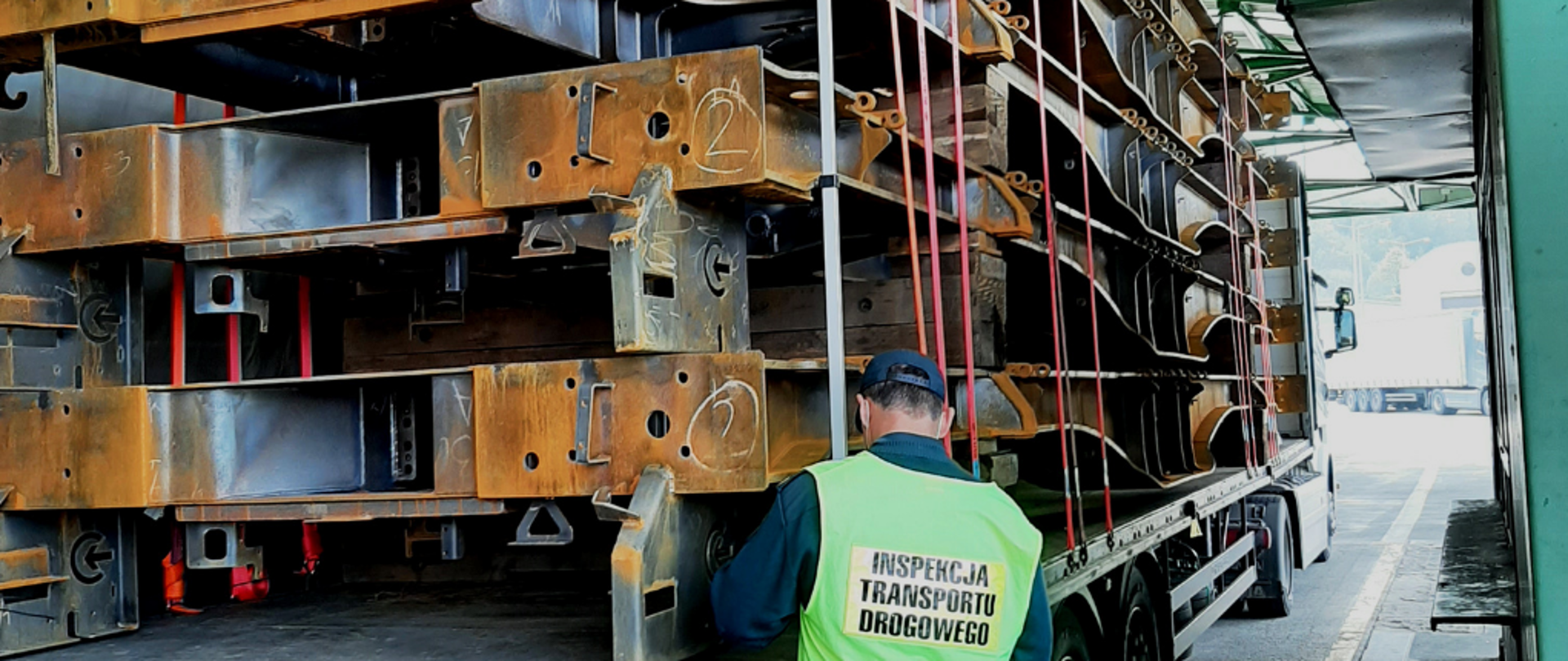 Inspektor ITD stoi obok naczepy, na której są stalowe ramy do wagonów. Przy pomocy wysokościomierza sprawdza wysokość zestawu wraz z ładunkiem.