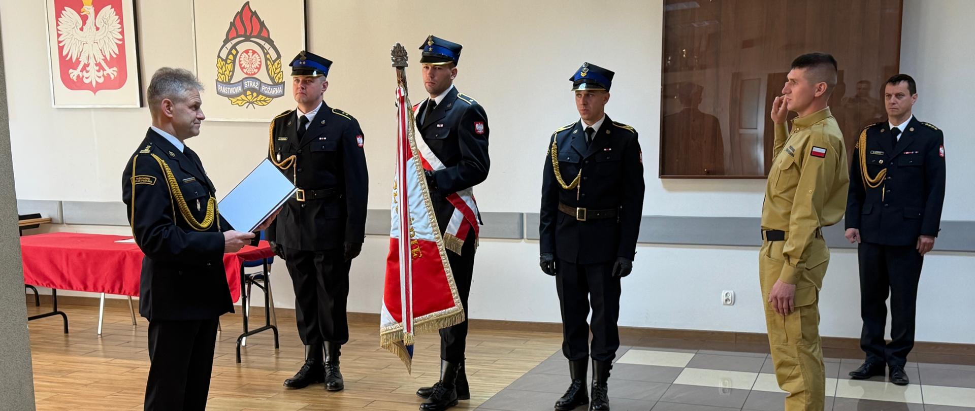 Uroczystość w komendzie Państwowej Straży Pożarnej. Na pierwszym planie strażak w mundurze służbowym składa ślubowanie przed komendantem, który odczytuje rotę z dokumentu. Obok stoją funkcjonariusze w galowych mundurach, z pocztem sztandarowym PSP. W tle widać godło Polski oraz emblemat Państwowej Straży Pożarnej.