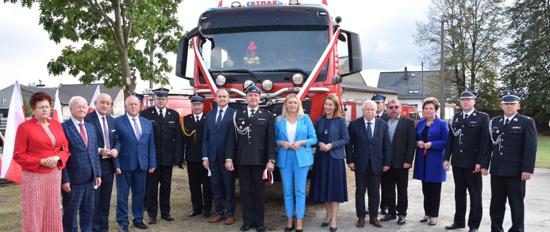 zdjęcie grupowe strażaków i osób cywilnych przed samochodem strażackiim w tle drzewa