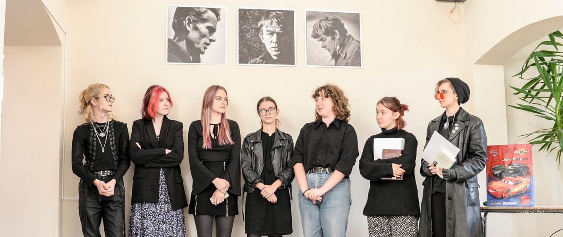 Zdjęcie siedmiorga uczniów przedstawiający skład obecnego i byłego samorządu uczniowskiego na tle beżowej ściany w holu szkoły. Nad głowami uczniów zawieszone na ścianie trzy czarnobiałe zdjęcia Władysława Hasiora en face w środku i z obu profili po bokach.