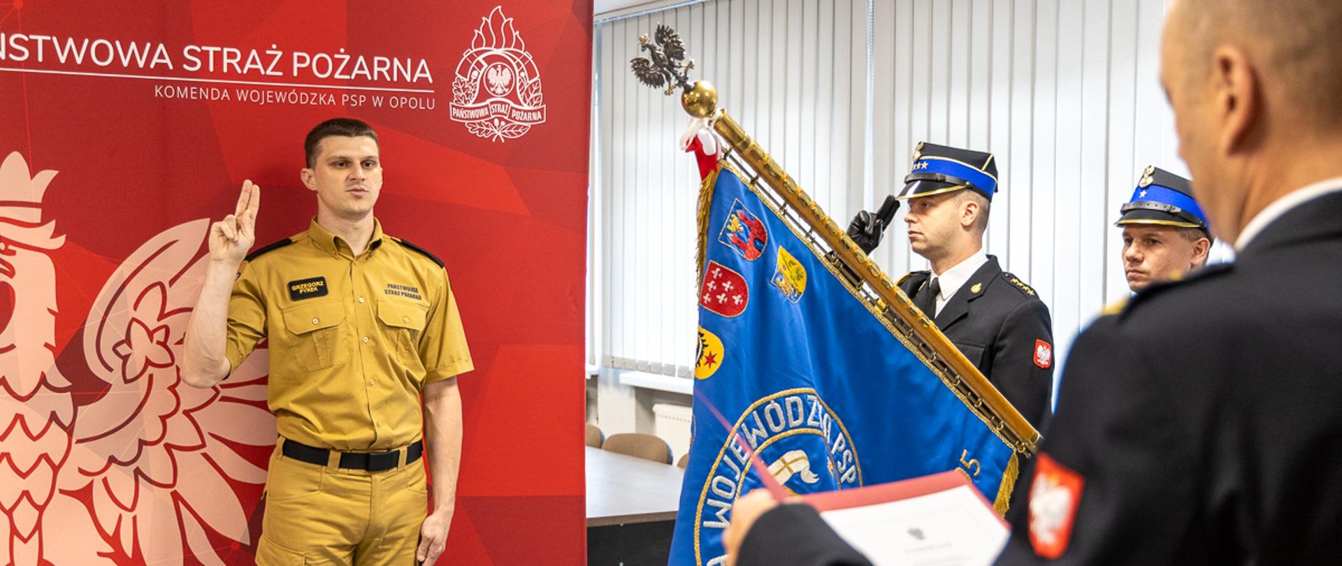 Po lewej stronie kadru stoi str. Grzegorz Pyrek, uniesioną dłonią składając ślubowanie. Po prawej stronie widoczna jest grupa oficerów PSP w granatowych mundurach galowych, trzymających sztandar wojewódzki PSP. Jeden z oficerów czyta tekst roty z dokumentu. W tle znajdują się pionowe żaluzje okienne, a całość odbywa się w sali odpraw Komendy Wojewódzkiej PSP w Opolu. Zdjęcie ukazuje kluczowy moment uroczystości — wypowiedzenie słów ślubowania.