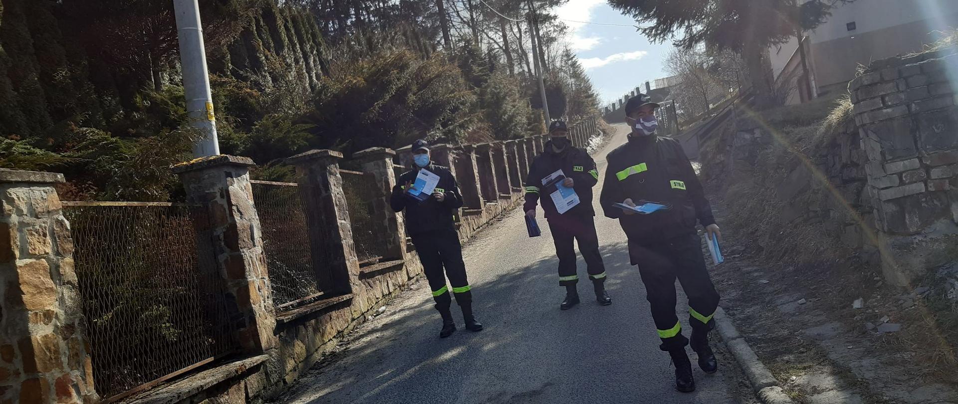 Zdjęcie przedstawia trzech strażaków Ochotniczej Straży Pożarnej z Manasterca trzymających ulotki dotyczące szczepień przeciwko COVID-19. Strażacy dostarczają ulotki mieszkańcom gminy lesko.