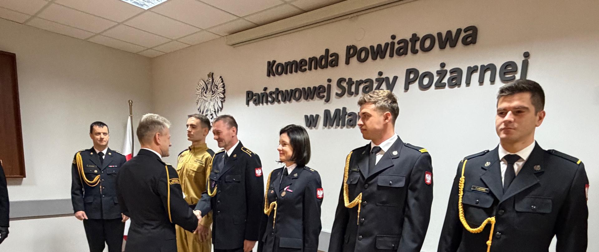 Uroczystość w Komendzie Powiatowej PSP w Mławie. W sali stoi rząd strażaków w galowych mundurach. Oficer w czarnym mundurze podaje rękę jednemu z funkcjonariuszy, jakby gratulował mu lub wręczał nominację. Obok stoją kolejni strażacy, w tym jeden w ubraniu koszarowym koloru żółtego. Na ścianie widoczny jest napis „Komenda Powiatowa Państwowej Straży Pożarnej w Mławie” oraz godło Polski.