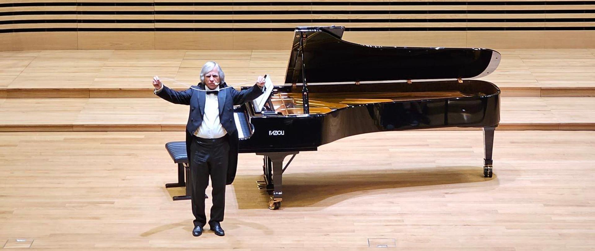 Mężczyzna we fraku stoi przed fortepianem Fazioli na estradzie sali koncertowej trzymając w wysoko podniesionych dłoniach biały sznurek. Z przodu widać od tyłu fragment widowni z publicznością.