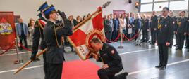 Na zdjęciu widoczna jest uroczysta ceremonia w jednostce Państwowej Straży Pożarnej. Strażak w galowym mundurze klęczy na czerwonym dywanie i całuje sztandar, który trzymają inni strażacy. Jeden z nich oddaje honory przez salut. W tle zgromadzeni są goście i funkcjonariusze, obserwujący wydarzenie. Całość odbywa się w hali z otwartą bramą garażową, a przestrzeń oddzielona jest czerwonymi barierkami.