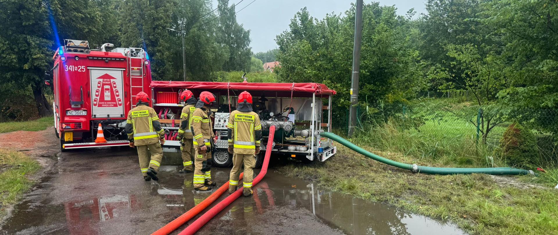 Strażacy stojący przy samochodzie pożarniczym. Pompuja wodę z zalanych terenów. 