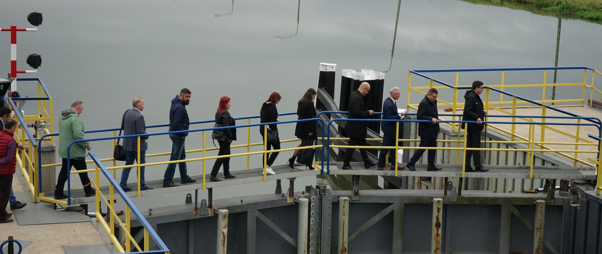 Zdjęcie przedstawia fragment infrastruktury wodnej - jaz we Wróblinie. Na zdjęciu widać tamę, nad którą jest pomost. Przez pomost przechodzą ludzie, oglądający inwestycję. W tle widać rzekę Odrę. 