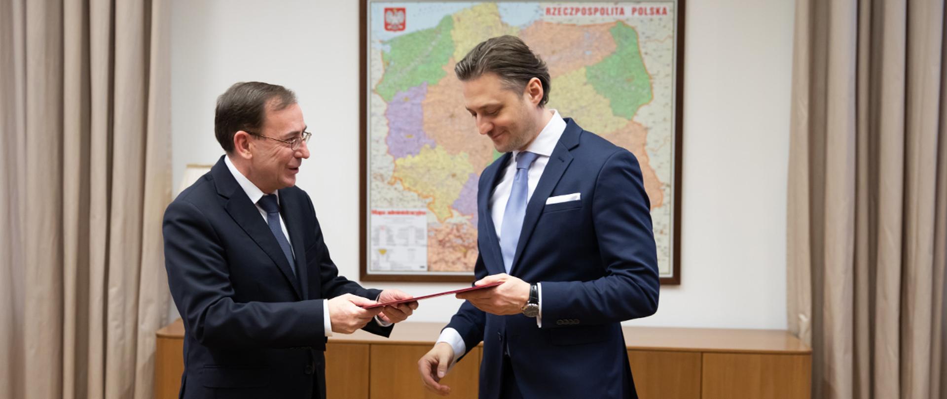 Na zdjęciu widać ministra Mariusza Kamińskiego wręczającego akt powołania na stanowisko wiceministra Bartoszowi Grodeckiemu. W tle widać mapę administracyjną Polski.