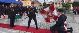 Rynek miasta na którym trwają uroczystości nadania sztandaru dla Komendy Powiatowej PSP w Gostyniu. Na pierwszym planie czerwony dywan na którym stoi Zastępca Komendanta Głównego PSP nadbryg. Arkadiusz Przybyła oraz Komendant Powiatowy ze sztandarem przed, którym klęczy poczet sztandarowy. 