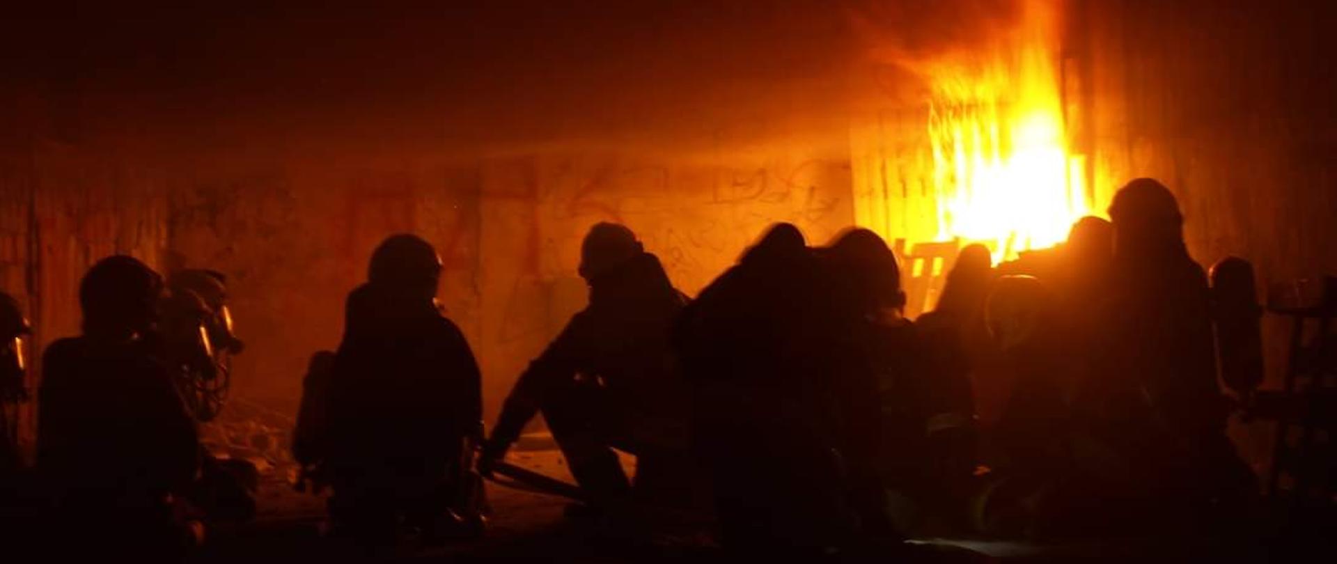Zdjęcia przedstawia strażaków ubranych w ubranie specjalne znajdujących się pomieszczeniu w którym jest pożar oraz silne zadymienie.