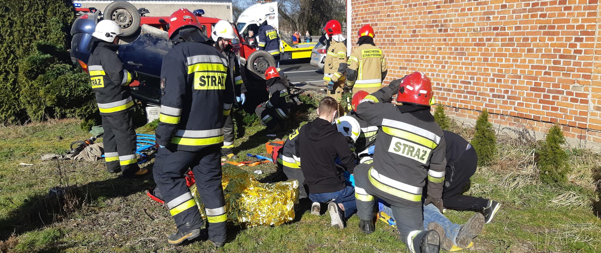 Zdjęcie przedstawia strażaków udzielających pomocy osobie poszkodowanej w wypadku samochodowym, w tle leżący na dachu samochód osobowy i budynki mieszkalne