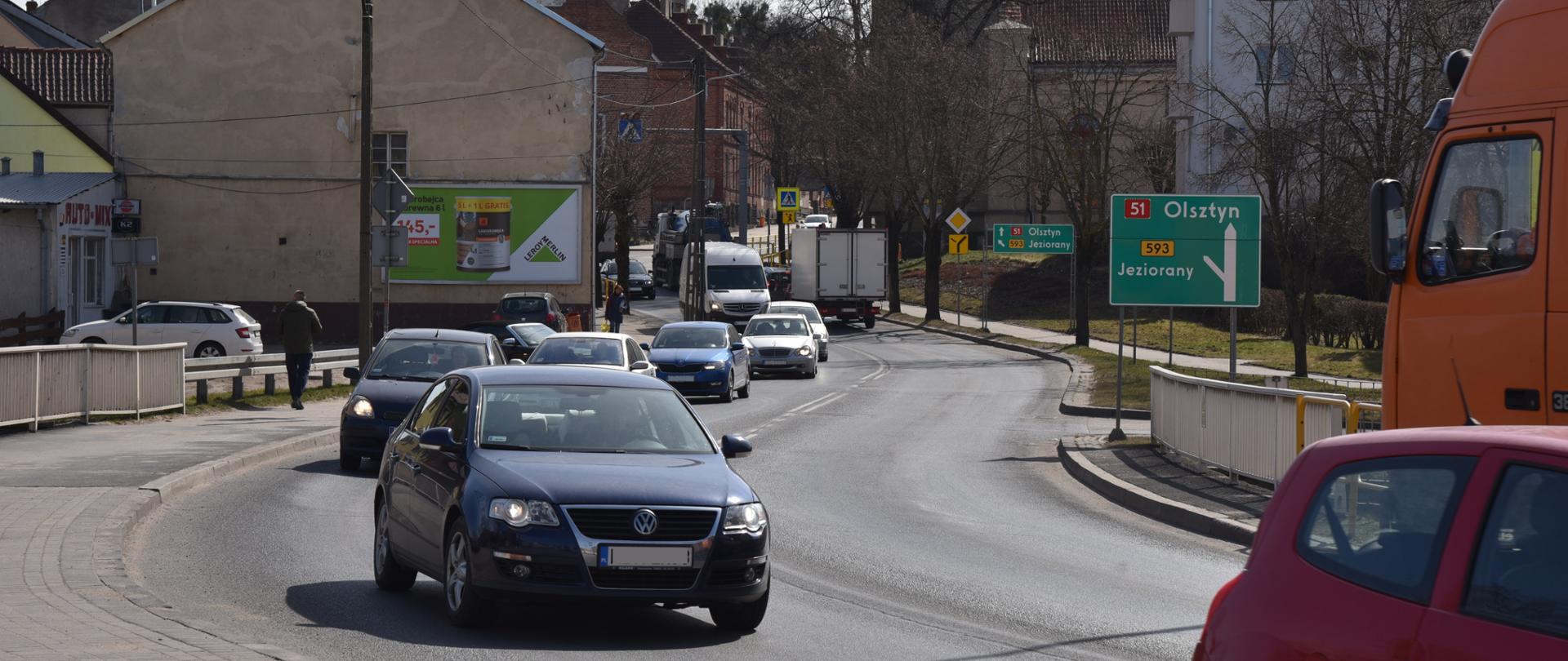 Ulica w centrum miasta. Wokół zabudowa mieszkaniowa, w tle wieża kościoła. Na ulicy duży ruch samochodów.