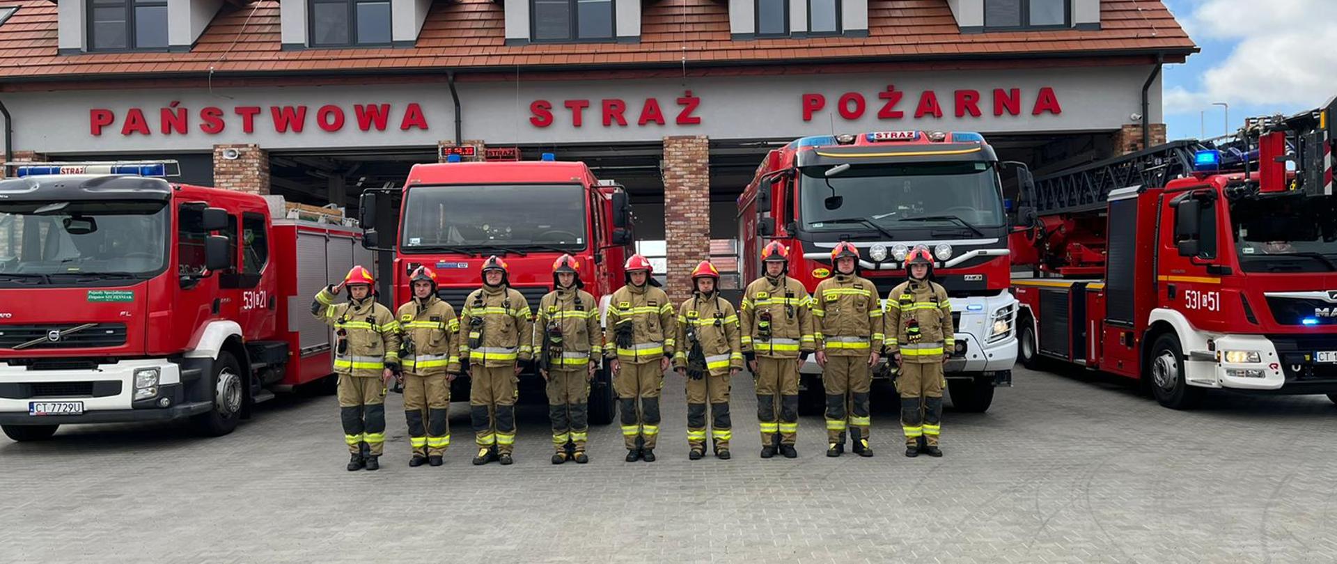 Przed budynkiem Komendy Powiatowej Państwowej Straży Pożarnej w Świeciu stoją cztery samochody ratowniczo-gaśnicze. Przed nimi w szeregu stoi dziewięciu funkcjonariuszy PSP ubranych w ubrania specjalne w hełmach. Dowódca zmiany stojący pierwszy z lewej oddaje honor. 