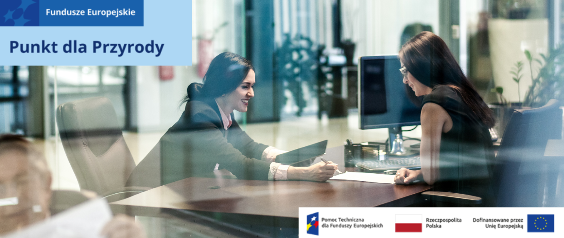 Dwie uśmiechnięte kobiety, siedzące na przeciwko siebie nad dokumentami leżącymi na stole biurowym. Jedna z nich wskazuje drugiej coś długopisem. 