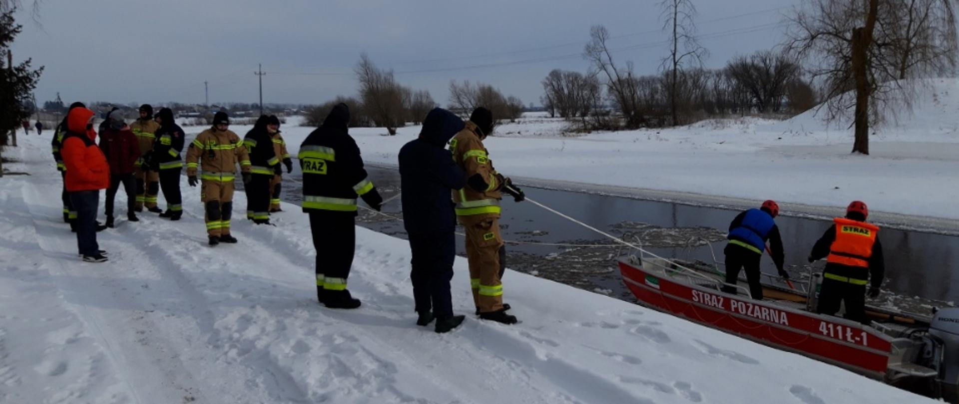 Dwóch strażaków ubranych w kamizelki ratunkowe na łodzi prowadzących działania poszukiwawcze na rzece Nurzec. Na brzegu pozostali strażacy biorący udział w akcji.