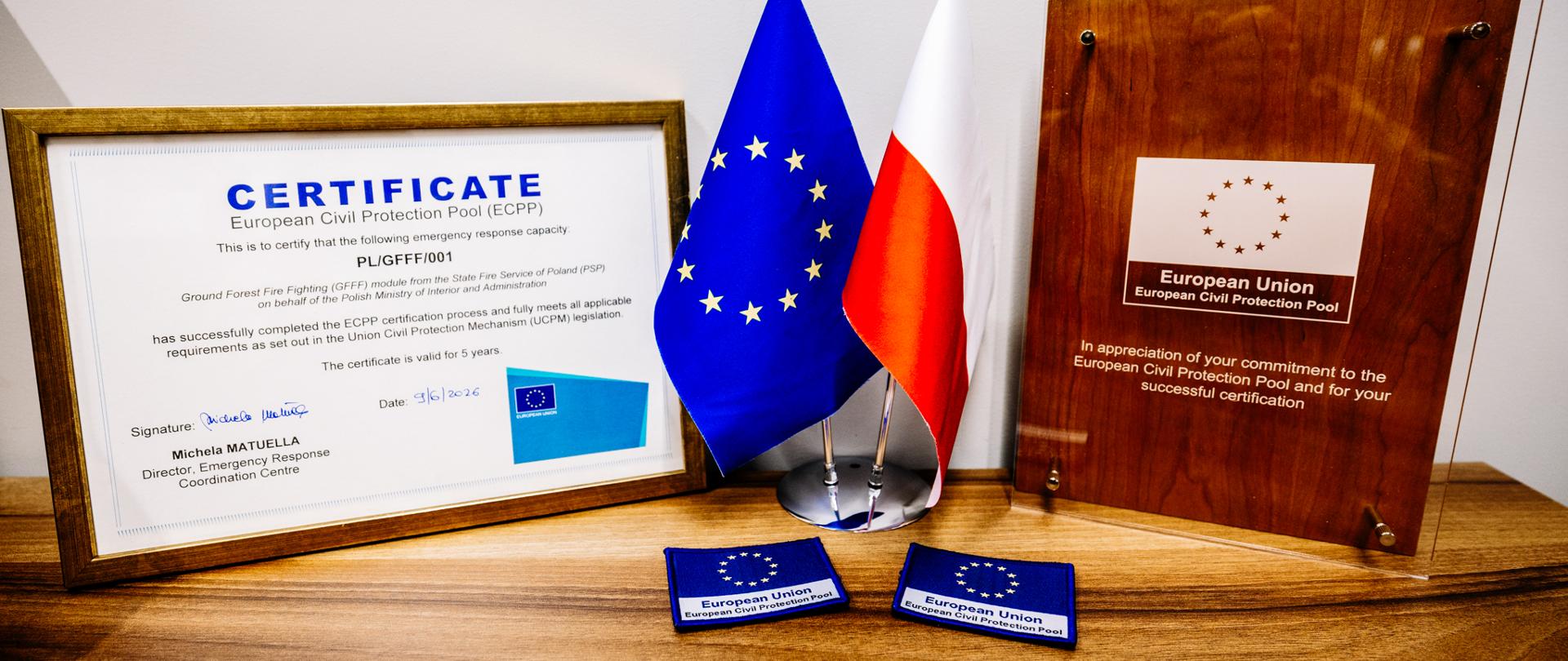 Certyfikat European Civil Protection Pool dla modułu GFFF Poland ustawiony obok flag Unii Europejskiej i Polski. Po prawej stronie widoczna jest pamiątkowa tablica Europejskiej Puli Ochrony Ludności, a na pierwszym planie dwie naszywki z symbolem Unii Europejskiej.
