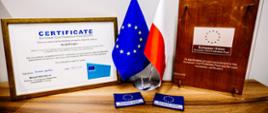 Certyfikat European Civil Protection Pool dla modułu GFFF Poland ustawiony obok flag Unii Europejskiej i Polski. Po prawej stronie widoczna jest pamiątkowa tablica Europejskiej Puli Ochrony Ludności, a na pierwszym planie dwie naszywki z symbolem Unii Europejskiej.