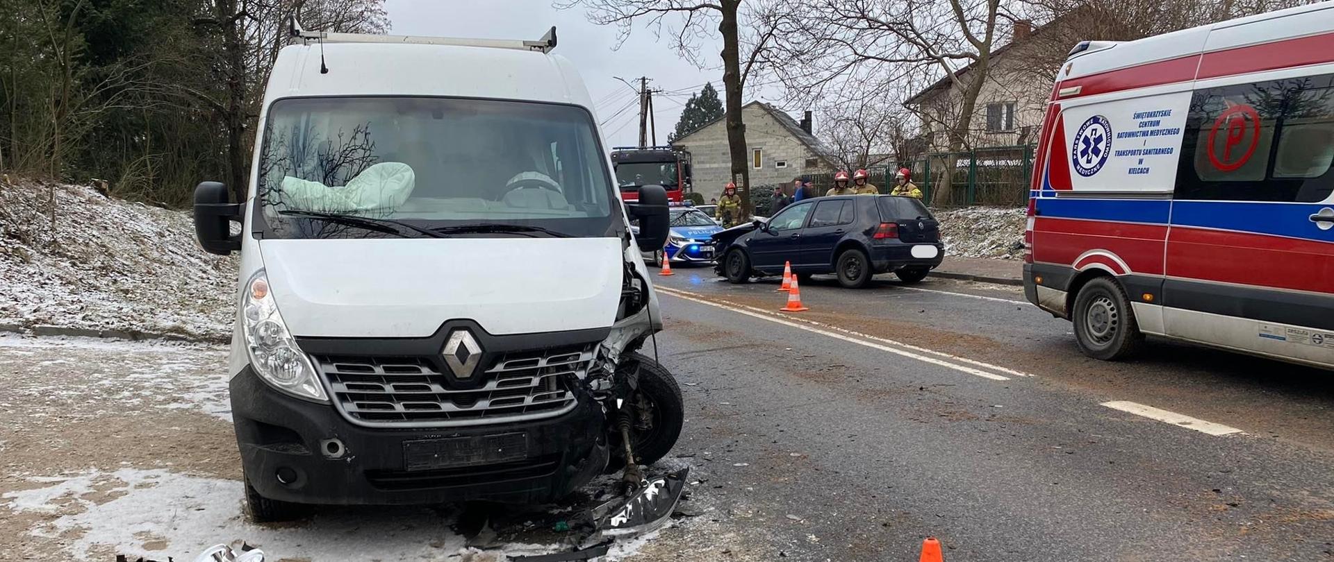 Na zdjęciu przedstawiono: na pierwszym planie samochód Renault, który uległ kolizji i stoi na poboczu ze zniszczonym lewym nadkolem. Po prawej stronie na ulicy stoi ambulans Ratownictwa Medycznego, w głębi jest zniszczony granatowy Volkswagen, a za nim pojazdy pozostałych służby: radiowóz i samochód pożarniczy.