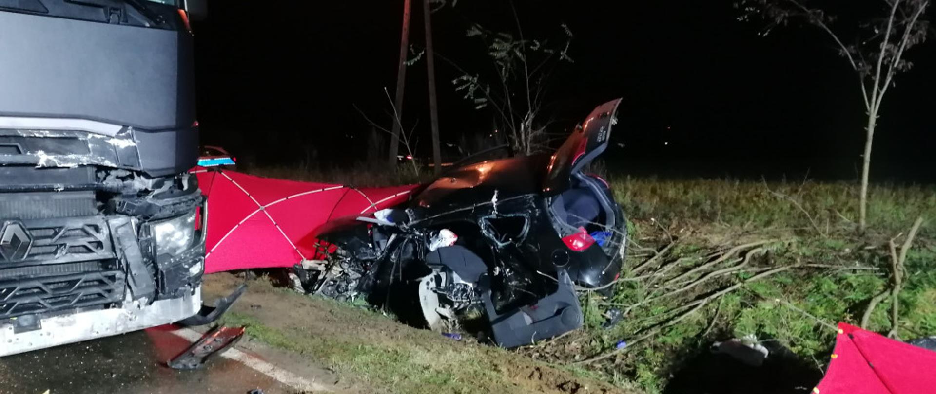 Widać ciemny samochód ciężarowy z uszkodzonym przodem, w rowie widać uszkodzony ciemny samochód osobowy