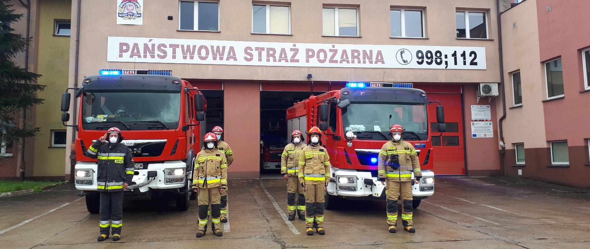 Widoczny budynek komendy psp w Hajnówce, dwa wozy gaśnicze wraz z ratownikami stojącymi na baczność, włączone sygnały świetlne