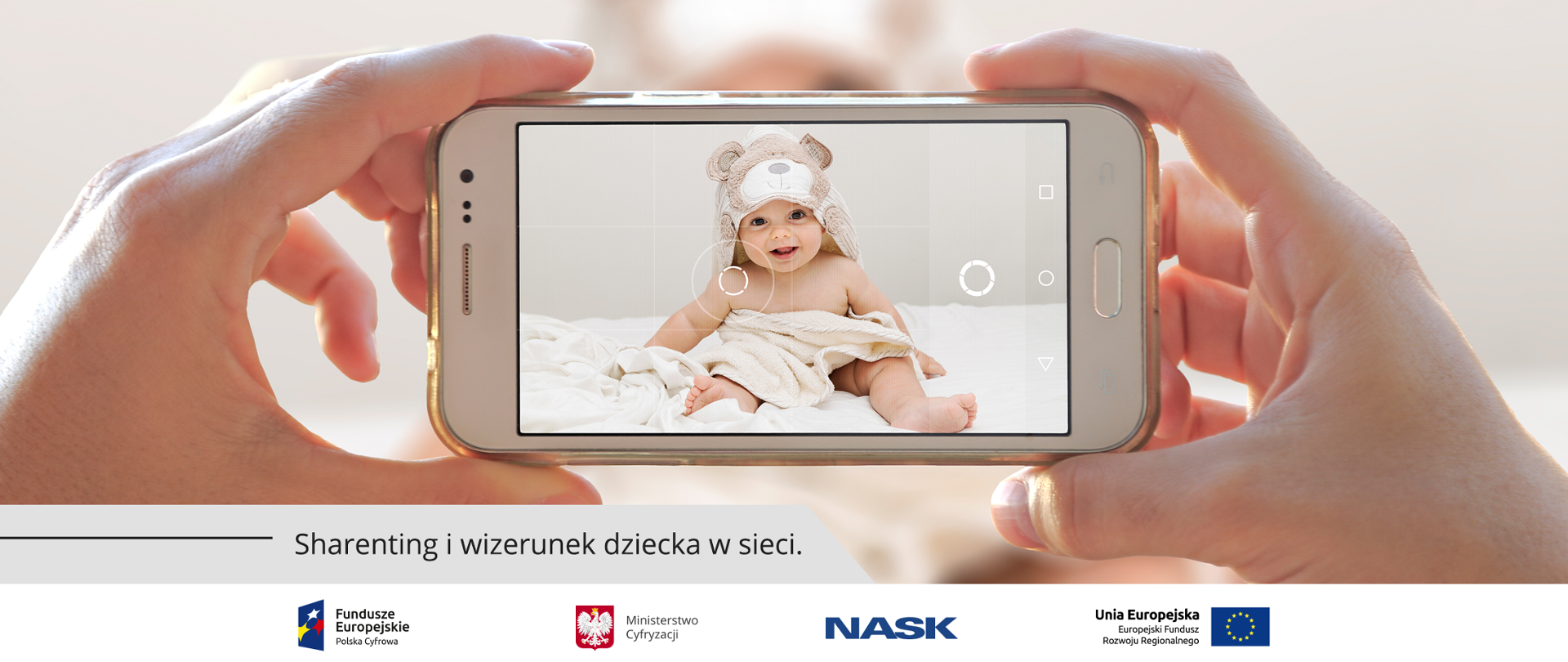 Bliskie ujęcie dłoni trzymających w poziomie telefon komórkowy podczas robienia zdjęcia. Na ekranie widać siedzącego na łóżku niemowlaka po kąpieli. Dziecko przykryte jest ręcznikiem, na głowie ma kaptur w kształcie misia. W lewym dolnym rogu obrazka napis: Sharenting i wizerunek dziecka w sieci.