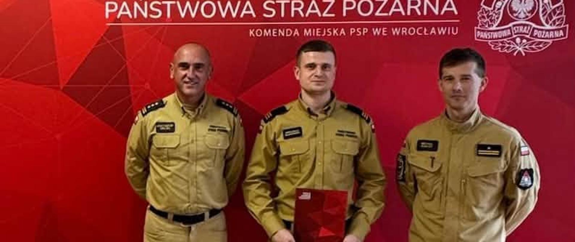 Trzech strażaków w beżowo-piaskowych mundurach stoi w pomieszczeniu na tle czerwonej ścianki prasowej z napisem „Państwowa Straż Pożarna Komenda Miejska PSP we Wrocławiu”. Mężczyzna stojący w środku trzyma w rękach czerwoną teczkę