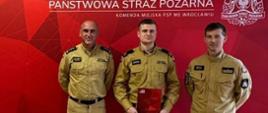 Trzech strażaków w beżowo-piaskowych mundurach stoi w pomieszczeniu na tle czerwonej ścianki prasowej z napisem „Państwowa Straż Pożarna Komenda Miejska PSP we Wrocławiu”. Mężczyzna stojący w środku trzyma w rękach czerwoną teczkę
