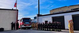 Na zdjęciu moment uroczystego wciągnięcia na maszt flagi państwowej, przy maszcie poczet sztandarowy, strażacy ustawieni w szeregu , w tle samochód pożarniczy, podniosły nastrój 