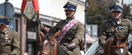 Żołnierze podczas jazdy na koniach. Jeden z żołnierzy trzyma w ręku sztandar. 