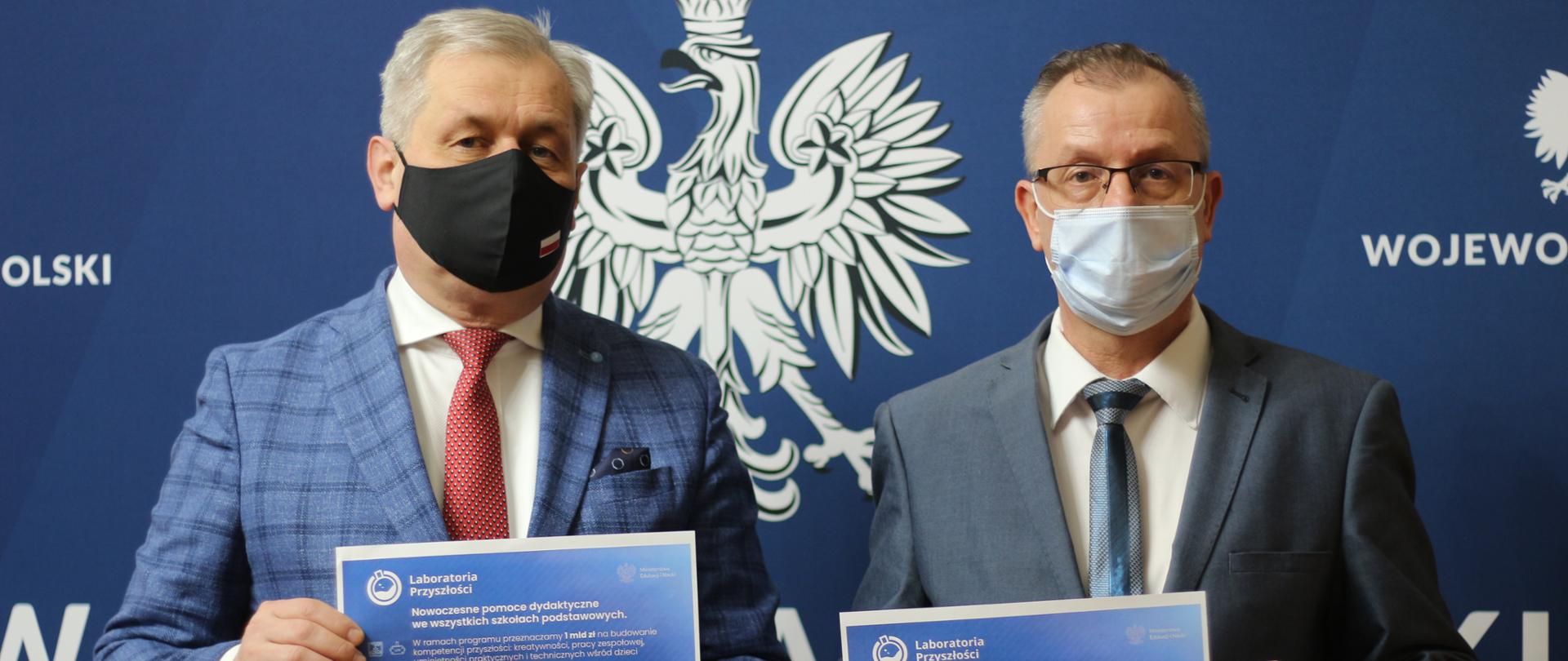 Na zdjęciu wojewoda opolski oraz wicekurator oświaty. W dłoniach trzymają plakaty z informacjami dot. wartości dofinansowania dla opolskich szkół. W tle ścianka z logo Wojewody Opolskiego. 