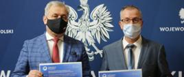 Na zdjęciu wojewoda opolski oraz wicekurator oświaty. W dłoniach trzymają plakaty z informacjami dot. wartości dofinansowania dla opolskich szkół. W tle ścianka z logo Wojewody Opolskiego. 