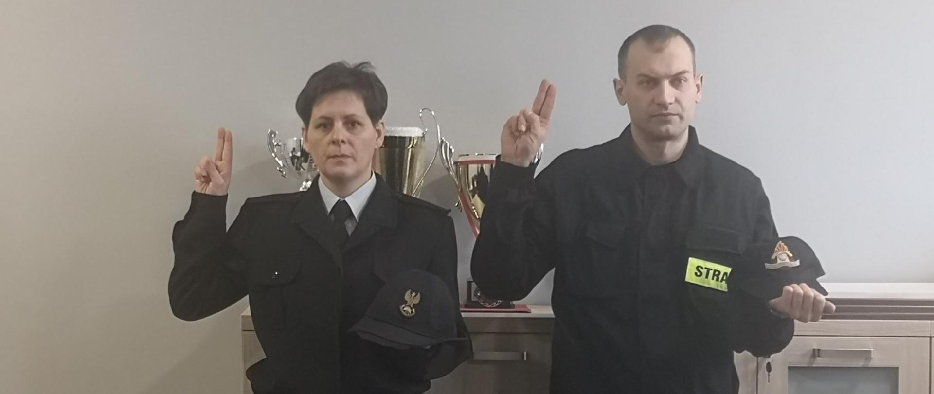 Na zdjęciu znajduje się 2 strażaków: mężczyzna w umundurowaniu koszarowym koloru czarnego oraz kobieta w umundurowaniu galowym koloru czarnego. Strażacy mają podniesione prawe dłonie do góry w geście ślubowania, natomiast na lewych dłoniach mają położone nakrycia głowy.