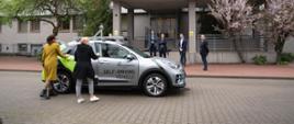 Przed wejściem do szarego budynku z napisem Łukasiewicz - Przemysłowy Instytut Motoryzacji stoi szary samochód z napisem na drzwiach Self-Driving vehicle, obok niego stoi wiceminister Mrówczyńska i kobieta w żółtym płaszczu.
