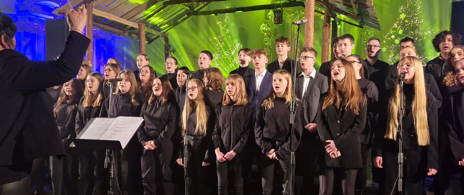 Zdjęcie przedstawia śpiew chórzystów podczas występu w kościele. Chórzyści ubrani są na czarno i ustawieni w kilku rzędach. Z nad ich głów widać dwuspadowy dach szkopki betlejemskiej. Tło stanowią choinki i oświetlenie w kolorze zielonym i niebieskim. Przed chórem ustawione są mikrofony, a przed nimi stoi czarny pulpit z nutami. Z lewej strony widać fragment dyrygenta z uniesioną do góry dłonią trzymającą batutę. 