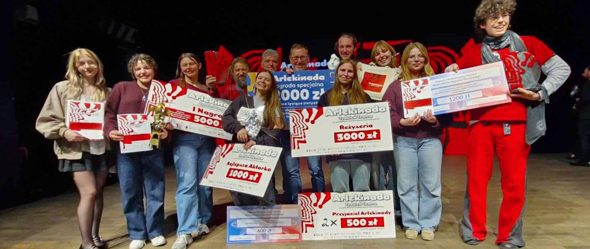 Na scenie teatralne stoi kilkanaście roześmianych osób - siedem dziewcząt, dwóch chłopców i trzech mężczyzn. Wszyscy trzymają w dłoniach duże dyplomy i informacje o zdobytych nagrodach Arlekinady. Dyplomy są na białym tle, z lewej strony czerwona grafika przypominająca głowę, na górze napis ARLEKINADA TYDZIEŃ TEATRU, niżej czerwone informacje: najlepsza aktorka, reżyser, I nagroda, Przyjaciel Arlekinady oraz przyznane kwoty pieniężne. Mężczyzna z tyłu trzyma niebiesko- żółtą informację o nagrodzie pieniężnej.