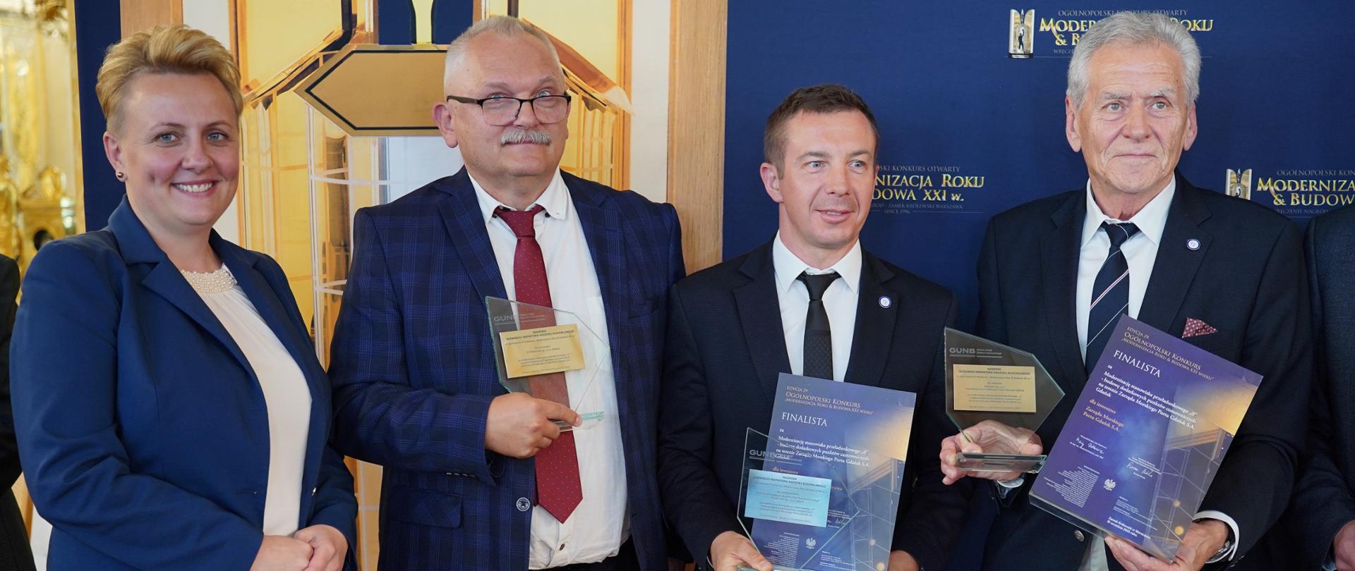 Laureaci i reprezentantka GUNB przed ścianką z logo konkursu „Modernizacja Roku & Budowa XXI wieku”. Trzech mężczyzn trzyma w rękach statuetki i dyplomy finalisty. Wszyscy pozują do zdjęcia po odebraniu nagrody.