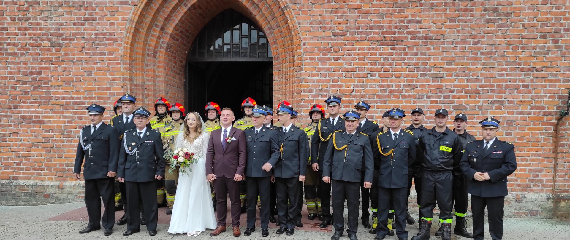 Przed kościołem stoi para młoda ze strażakami. Strażacy w mundurach wyjściowych i bojowych.