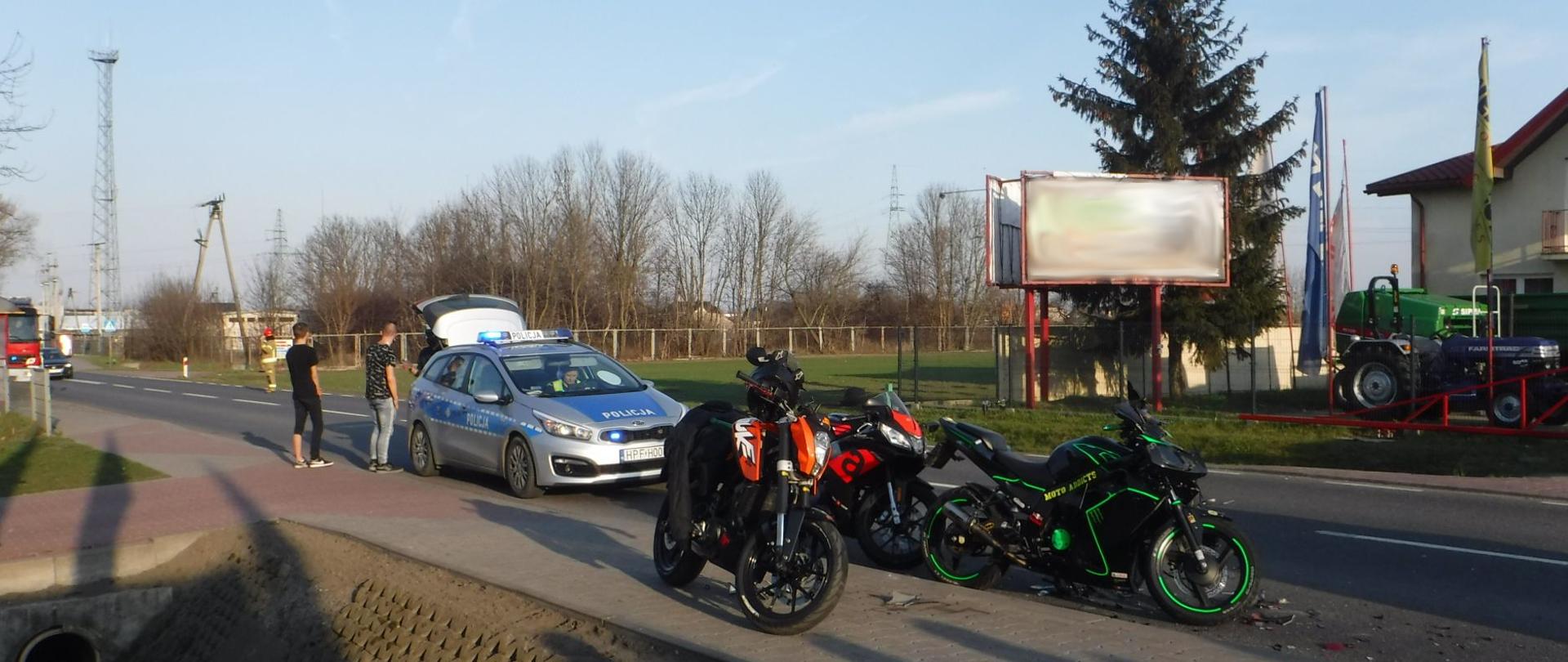 Na zdjęciu widać trzy motocykle, radiowóz, dwóch uczestników zdarzenia, czerwony samochód strażacki oraz strażaka kierującego ruchem na drodze. Strażak ubrany jest w żółte ubranie oraz czerwony hełm. W tle widać maszyny rolnicze u sprzedawcy oraz drzewa.