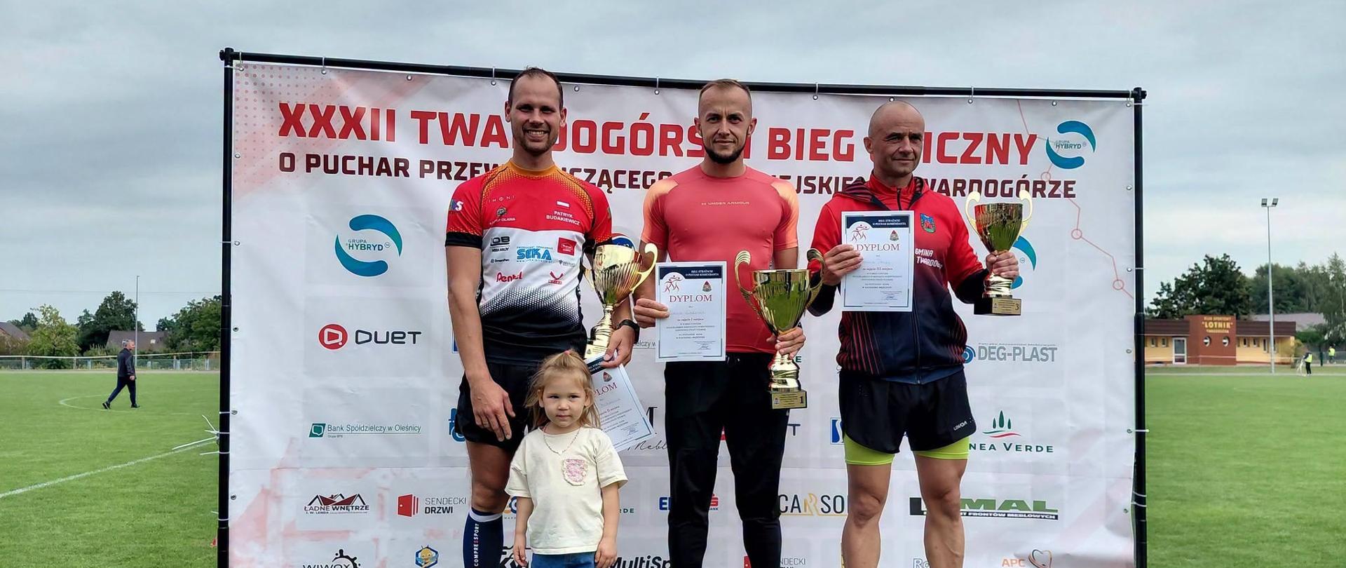 Zdjęcie przedstawia trzech mężczyzn stojących na podium podczas dekoracji zwycięzców XXXII Twardogórskiego Biegu Ulicznego o Puchar Przewodniczącego Rady Miejskiej Twardogóry. 