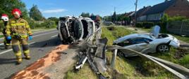 Zdjęcie przedstawia samochód osobowy po wypadku samochodowym, który wjechał w zaparkowany przy drodze inny pojazd. Jeden z nich wylądował w rowie, a drugi leży na boku na asfalcie. Z pojazdu wyciekły płyny eksploatacyjne. Obok pojazdu chodzą strażacy. 