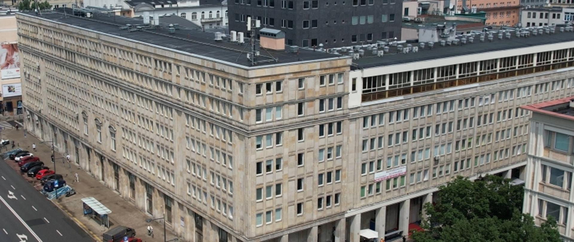 Wielopiętrowy budynek o modernistycznej architekturze przy ulicy z samochodami i oznaczeniami drogowymi.
