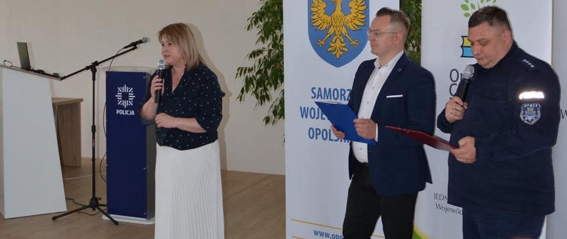 W wysokiej auli kobieta w czarnej bluzce i białej spódnicy przemawia z mikrofonem w ręku. Obok stoi mężczyzna w garniturze, a dalej mężczyzna w uniformie policyjnym.