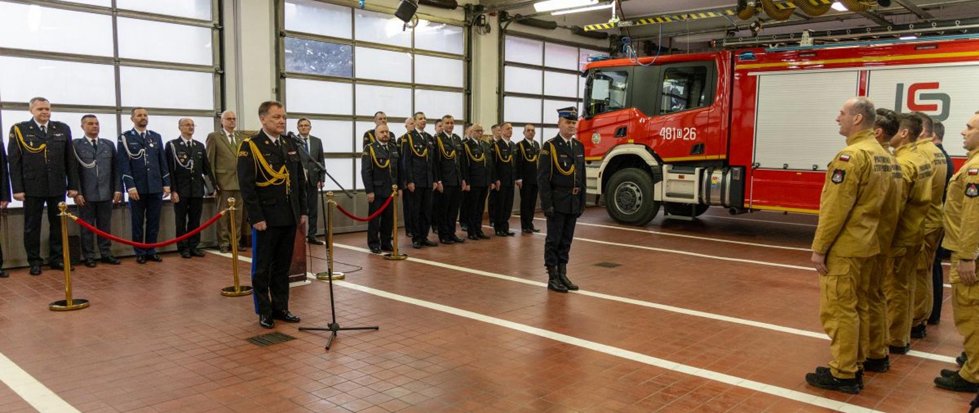 Uroczystość w garażu Komendy Powiatowej PSP – przemówienie przy mikrofonie, obecność funkcjonariuszy w galowych i bojowych mundurach oraz wóz strażacki w tle.