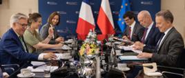 Spotkanie delegacji Polski i Francji przy długim stole konferencyjnym. Uczestnicy siedzą naprzeciw siebie, przeglądając dokumenty i prowadząc rozmowy. Na pierwszym planie widoczne są mikrofony, notatniki, filiżanki, szklanki oraz srebrne dzbanki. W centrum stołu znajduje się kompozycja z żółtych i różowych kwiatów. W tle ustawiono flagi Francji, Polski i Unii Europejskiej na tle granatowej ścianki z logotypami Ministerstwa Spraw Zagranicznych RP. Uczestnicy spotkania są ubrani w formalne stroje biznesowe, w tym garnitury, koszule, krawaty oraz marynarki.