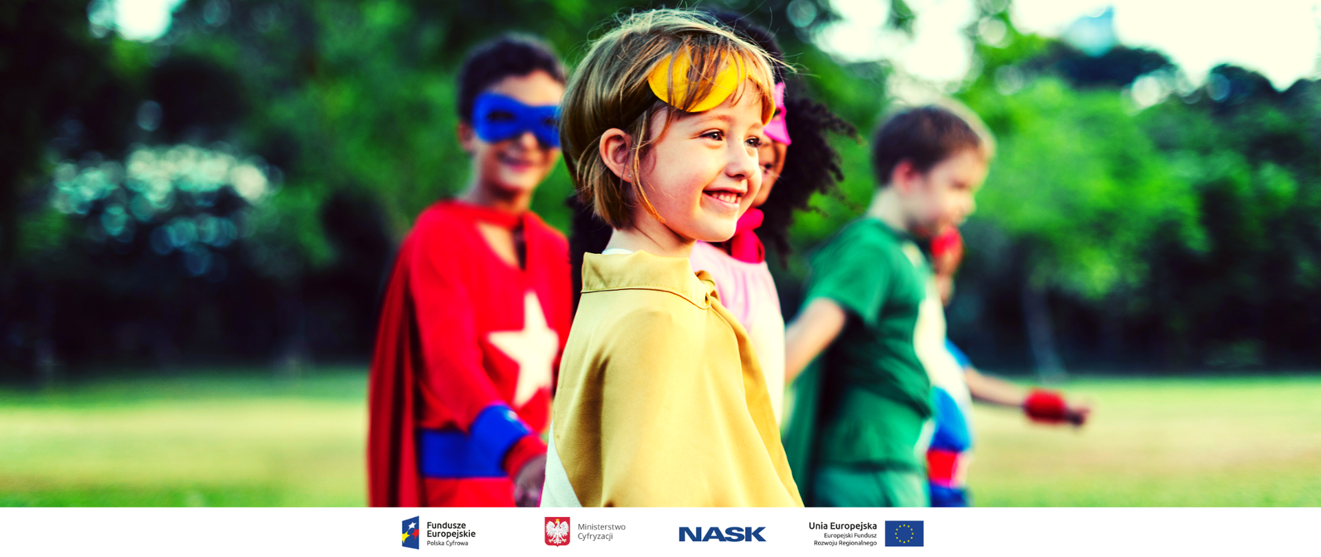 Kilkoro dzieci przebranych za bohaterów kreskówek. Bawią się w parku. Na pierwszym planie uśmiechnięta dziewczynka w żółtej pelerynie i z maską na czole. W tle nieostre drzewa.