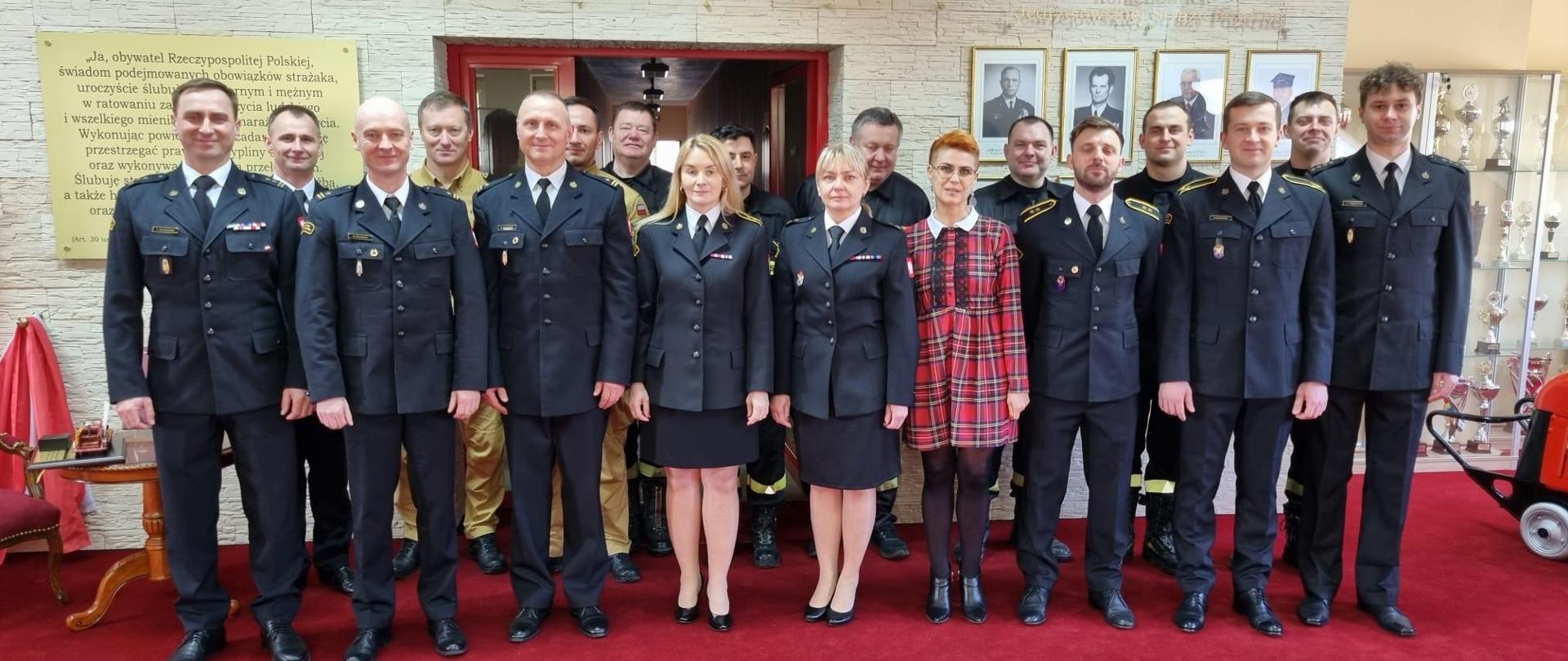 Funkcjonariusze i pracownicy Komendy Powiatowej Państwowej Straży Pożarnej w Jędrzejowie na uroczystej zbiórce pożegnania emeryta. Wszyscy znajdują się na sali tradycji.