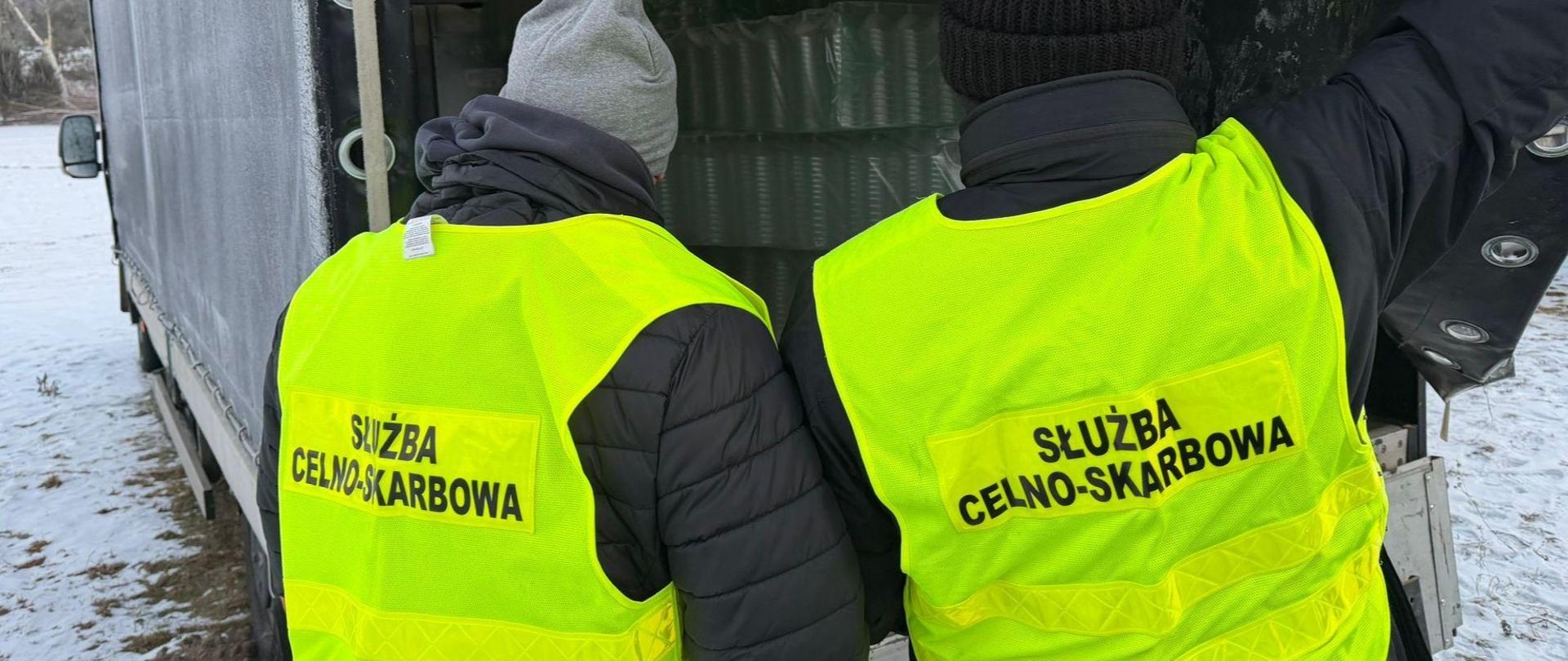 Dwie osoby w kamizelkach odblaskowych z oznaczeniem służby stoją tyłem przy otwartej ciężarówce, w której widać zapakowane pojemniki.