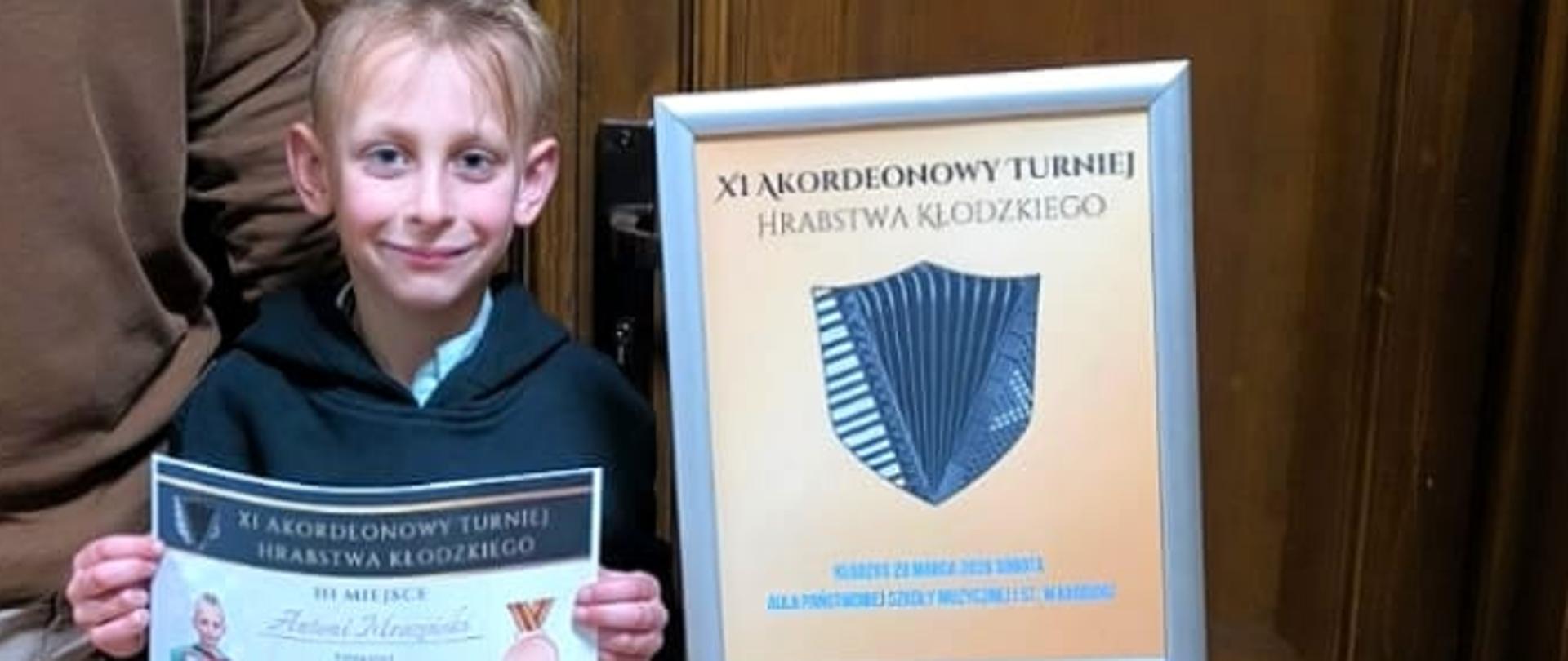 Zdjęcie przedstawiające Antoniego Mrozińskiego wraz ze swoim nauczycielem - panem Bogusławem Kłakiem przed salą koncertową w Kłodzku. Antek trzyma w rękach dyplom za zajęcie trzeciego miejsca w konkursie XI Akordeonowego Turnieju Hrabstwa Kłodzkiego.