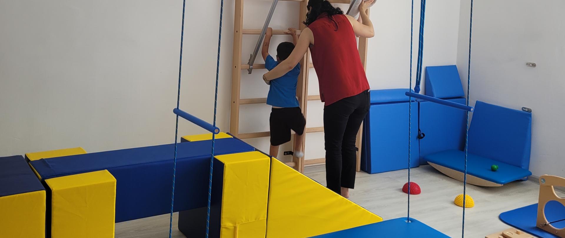 Sala terapeutyczna wyposażona w sprzęty pomocne do prowadzenia zajęć: maty, huśtawki, tory przeszkód, drabinki. Na drabinkę wspina się dwójka dzieci przy asekuracji terapeutki.