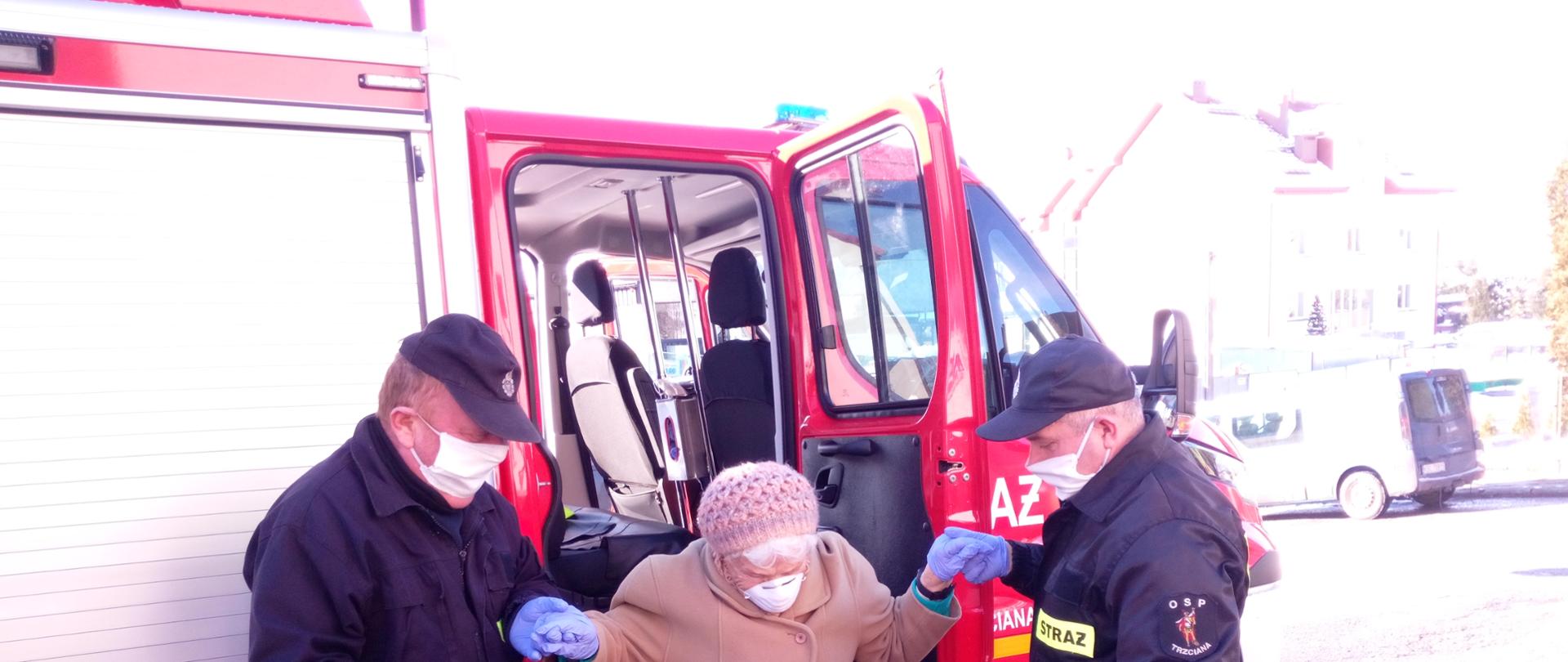 Pomoc przez strażaków Ochotniczej Straży Pożarnej w Trzcianie w wyjściu z pojazdu osobie starszej po dojeździe do punktu szczepień.